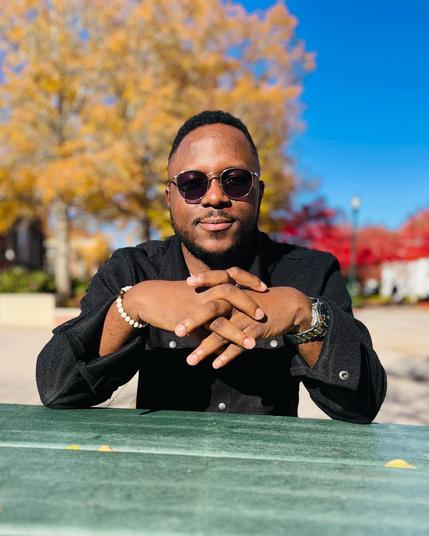 Head-and-shoulders color photo of Tobi Ogundiran, a Black man with short, black hair and a matching short, full beard and mustache. He wears a black button-up, a bracelet of white beads, a watch with a metal band, and metal-rimmed aviator sunglasses that are taking on enough sun that you can see his dark eyes through them. Trees are visible in the distance. He rests both elbows on a green picnic table in front of him and sits with his fingers laced together. He looks directly into the camera with serious eyes and a tiny smile.