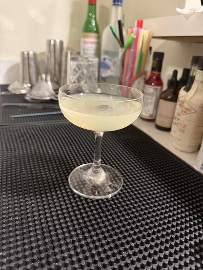 Slightly yellowish cocktail in a clear rounded coupe with a brandies cherry barely visible through the drink. Sitting on a bar mat with the tools and bitters of a home bar visible in the background.