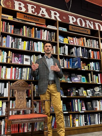 The authorNathaniel Ian Miller stands in front of shelves of books and below a sign with the name of the bookstore, Bear Pond Books. He is wearing a plaid wool blazer, and gold colored trousers.
