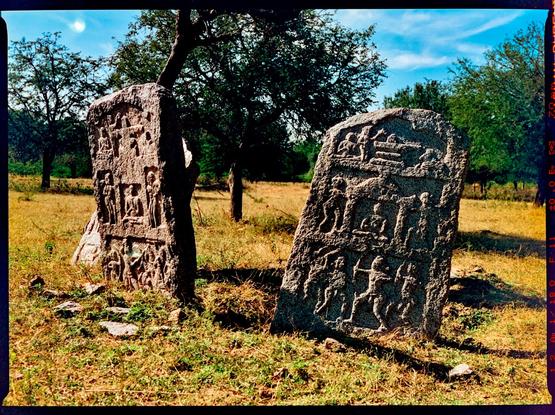 A color photo of two upright carved stones, in the shape of tombstones, showing 3 panels depicting the lives of people approximately 7-800 years ago.