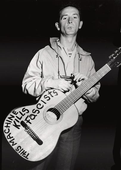 Woody Guthrie, a folk musician, with a harmonica on a string around his neck, poses with his acoustic guitar - the words written on the body of the guitar "This machine kills fascists".