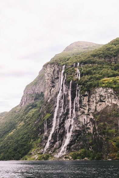 A stunning view of the Seven Sisters Waterfall in Norway, cascading down a steep, rocky mountainside surrounded by lush greenery. The waterfall plunges into a serene fjord, with overcast skies creating a dramatic and tranquil ambiance.