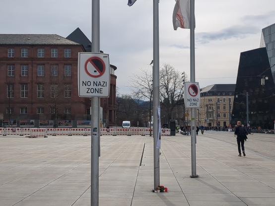 Fast menschenleerer Platz der Alten Synagoge in Freiburg. Im Vordergrund drei Fahnenmasten. An zwei Masten hängen Schilder mit der Aufschrift 'No Nazi Zone'