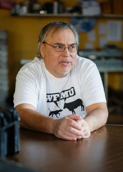 A man with salt-and-pepper hair and the beginnings of a salt-and-pepper beard sits at a table.  He wears wire-rimmed glasses and a white tee shirt bearing the Woof-Moo logo of radio station WFMU.  He appears to be engaged in a serious conversation.