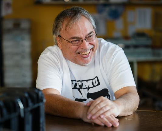 A man with salt-and-pepper hair and the beginnings of a salt-and-pepper beard, wearing wire-rimmed glasses and a white tee shirt bearing the Woof-Moo logo of radio station WFMU, leans on a tabletop and laughs.