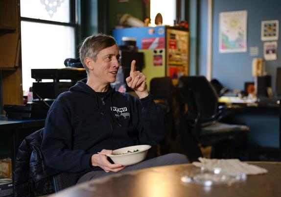 A man wearing a blue hoodie which bears the Woof-Moo logo of radio station WFMU holds a white bowl of food in one hand and holds up the index finger of the other hand as if making a point.