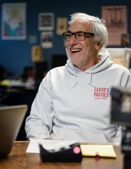A man with white hair and black-rimmed glasses laughs.  He wears a white hoodie bearing the legend "LEHTO'S /  PASTIES / Since 1947" in red.