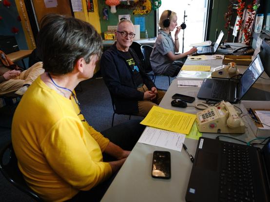 We see two phone volunteers and the emcee in a row at one long desk, and a bit of the lap of the third phone volunteer behind them.  The volunteer in the middle tilts his head and smiles quizzically (or enigmatically, or both) at the camera.