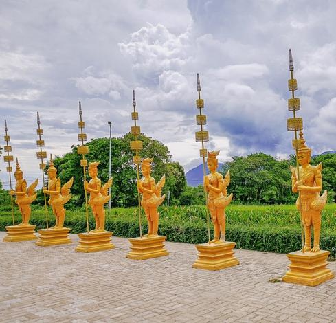 Photo shows a range of seven golden guardian figurines with golden spears in front of a gate to a tall mountain. The figurines have the upper body of a woman, and the lower part of a bird/swan. They carry the traditional headdress of supporting Apsara dancers. 
The skies are dark and stormy dramatic, and the mountain is covered by deep, green forest until the top covered in fog.