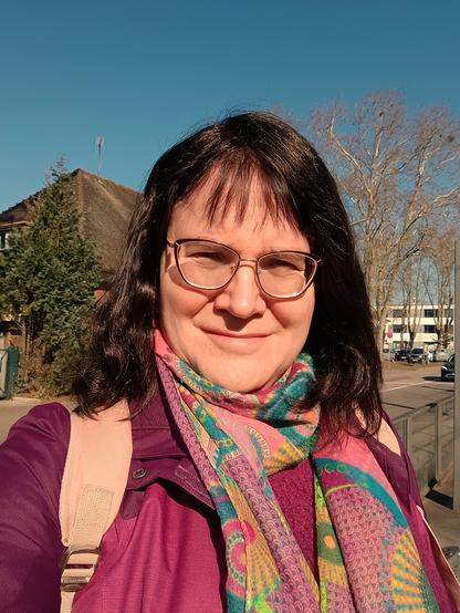 Ich, eine Frau mit schulterlangem braunem Haar in einer offenen Ponyfrisur, mit pink-grün gemustertem Schal, violettem Pullover und Outdoorjacke, an einer Tram-Haltestelle unter strahlend blauem Himmel.