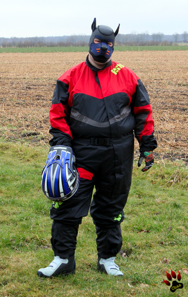 human pup wearing motocross raingear and mx boots standing outside looking into the camera, MX helmet resting on his leg