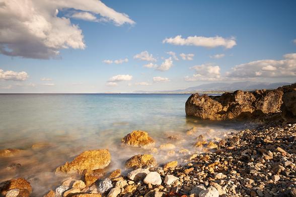 On the right side is the pebble beach with some rocks in the warm evening sun leading to the sea on the left and towards the horizon in the middle of the photo. The sea is milky due to the long exposure with light blue and turquoise tones. The sky above the sea is light blue with some larger white clouds.