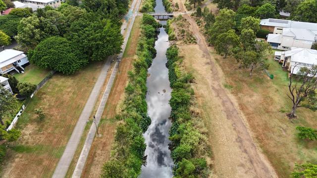 An ariel photo taken from directly above Kedron Brook, looking along its length. The brook is calm, and reflecting the clouds, with greenery on its banks. Pathways run along either side of it, with houses visible at the edge of the photo, their backyards opening towards the brook
