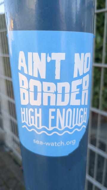 Adhesivo enganchado en una farola donde se lee "AIN'T NO BORDER HIGH ENOUGH - sea-watch.org