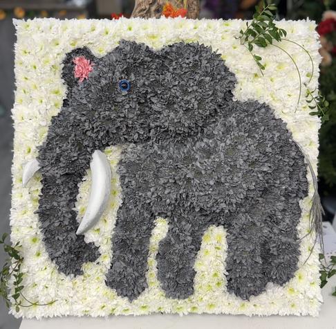 A 2 dimensional grey elephant, it's head turned slightly to the left, it's tail swishing, it's ears opened with tusks long and sharp, a trunk not far from the ground, looking for food stands against a white backdrop. This is not a real-life elephant though but one which has been created from flowers.
