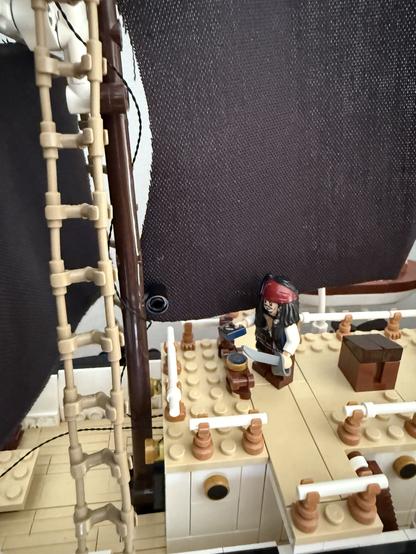 A close-up of a pirate-themed LEGO ship, featuring a miniature pirate figure with a sword standing on the deck near a chest and barrels, with a background of black sails and a ladder.