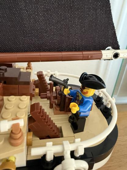 A pirate figure in a blue shirt and black hat stands on the deck of a small LEGO ship, holding a pistol. The ship features a dark sail, wooden steps, and various LEGO brick details.