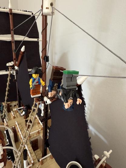 The image features a detailed model of a pirate ship, equipped with black sails and various rigging. Two Lego minifigures are prominently displayed: one is standing on a deck and the other is hanging upside down from the rigging. The ship