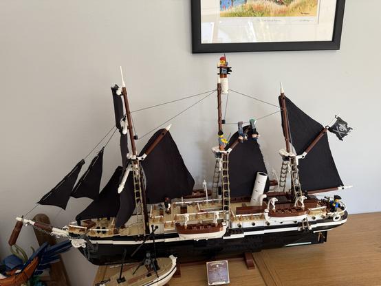 A detailed model of a pirate ship with black sails, featuring several miniature pirate figurines on deck and climbing the rigging. The ship is displayed on a wooden surface, with a decorative piece and a framed artwork visible in the background.