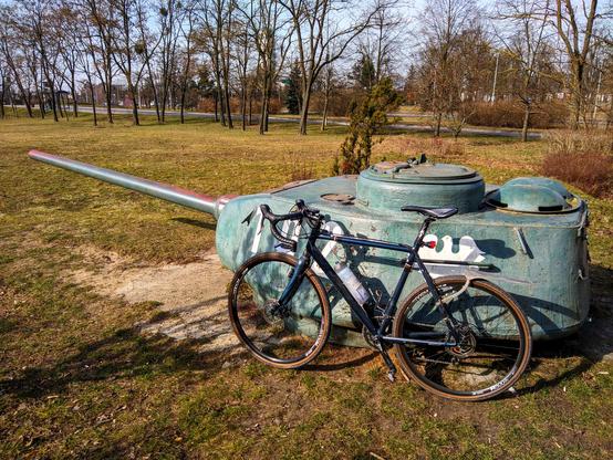 Ciemnoniebieski rower szutrowy Ridley stoi oparty o wieżyczkę czołgu T-35 z namalowanym numerem 102 i białym orłem, stojącą na trawniku. W tyle drzewa i ulica.