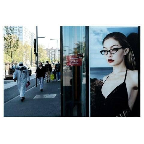 Une affiche publicitaire d’un mannequin au regard perçant et aux lunettes de style rétro occupe la moitié droite de l’image, reflétant la rue animée en arrière-plan. Sur le trottoir, des passants de divers horizons avancent, certains portant des masques ou des sacs de courses. La juxtaposition entre l’image figée et glamour du mannequin et le quotidien urbain crée un contraste saisissant entre idéal et réalité.