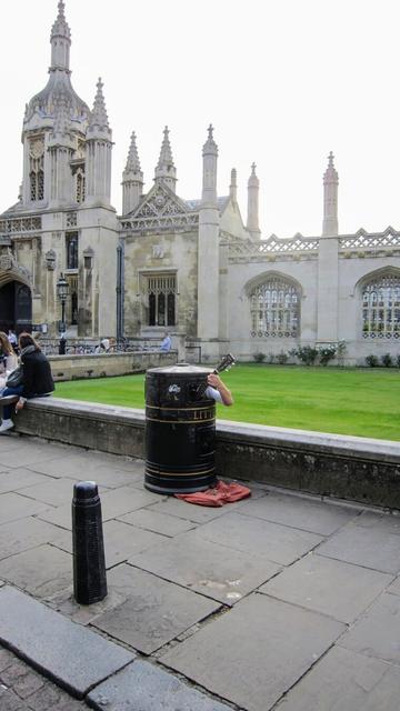 A street-level, slightly angled shot of a person playing a guitar while sitting inside a large, dark-coloured litter bin in front of King's College Chapel in Cambridge. The litter bin is positioned on a paved walkway, with a red cloth visible underneath for collecting money. The person's arm and guitar neck are the only parts of them visible, protruding from the dustbin. In the background, a grand, stone building with ornate architectural details is partially visible. The building, King's College Chapel, features tall spires and decorative windows. To the left, people are seated on a low wall, and a dark bollard is in the foreground.