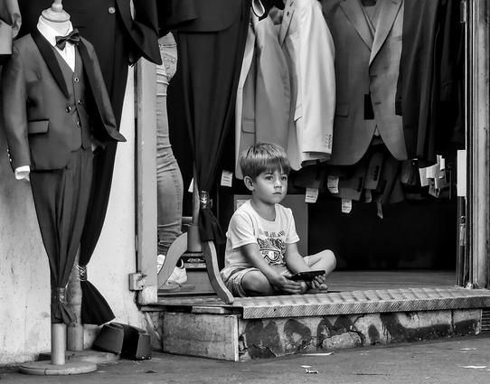 A little boy is sitting on the front step of a men's suit shop. He is staring into space. Next to him is a mannequin in a little boy's costume.