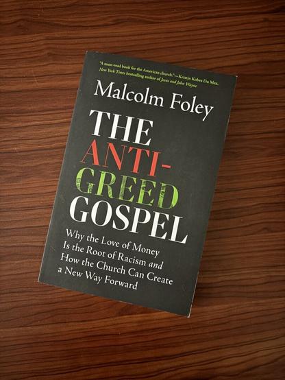 A copy of Dr. Foley’s “The Anti-Greed Gospel” rests on Caera’s desk. The book cover features the title in large red, white, and green text on a black background, and the green text has dollar print underlay.