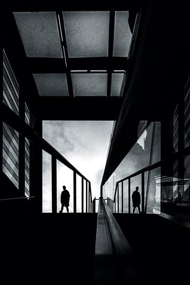 A lone individual walks up an escalator, flanked by their mirrored reflection on glass panels. The strong vertical and diagonal lines create depth, leading toward a bright opening that contrasts sharply with the surrounding shadows. The symmetry and reflections add a surreal, almost cinematic quality to the composition. (Photographed at Potsdamer Platz, Berlin, Germany.)