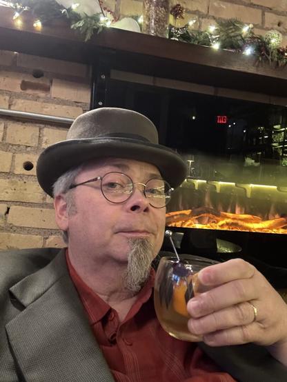 Selfie of a guy with a grey goatee toasting the camera with a cocktail. He is wearing a beaver top hat, glasses, and a grey suitcoat over and orange plaid shirt.