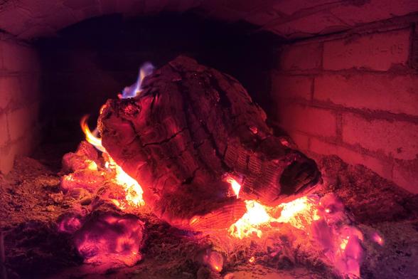 The image shows a burning log inside a wood-fired ove. The log is large and partially charred, glowing intensely with deep red and orange hues. The surface of the wood has deep cracks, revealing the embers inside.
There are smaller chunks of burning embers around the log, radiating a soft red and purple glow. A faint blue flame is visible near the top of the log, which often occurs when certain gases from the wood burn at high temperatures. The background consists of firebrick walls, which are slightly illuminated by the heat. The entire scene has a warm and rustic ambiance, reminiscent of traditional wood-burning ovens or fireplaces.