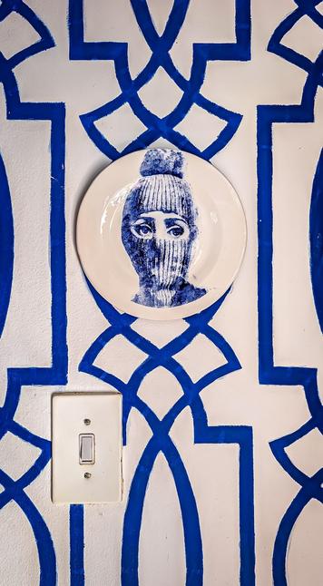 The image shows a decorative plate with a portrait of a person wearing a beanie or mask with only their eyes visible. The plate is mounted on a wall with blue and white geometric patterns. Below the plate, there is a light switch on the wall.
