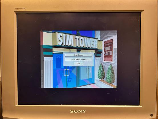 A photo of a flat-screen 4:3 Sony Bravia TV displaying the title screen for the game SimTower. There is a 3D render of a tower foyer with ‘SIM TOWER’ above the doors. There is a sign to the right saying ‘CREATED BY YOOT SAITO’. There is also a menu at the centre with options for New Tower, Load Saved Tower or Quit. L