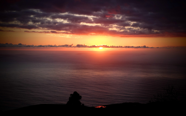 Blick von einem Berg in die Ferne. Die Sonne versinkt am Horizont und taucht den Himmel in warme Orange- und Rottöne. Dunkle Wolken ziehen darüber hinweg, während das Meer tief unten in sanften Wellen glitzert. Im Vordergrund sind silhouettenhafte Bäume und eine Hügelkette zu sehen.