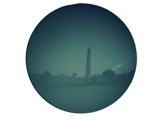 Greyey green roundel of an Irish round tower in the middle distance - a stone wall in the foreground - not fine detail as made with pinhole camera over three days this March
