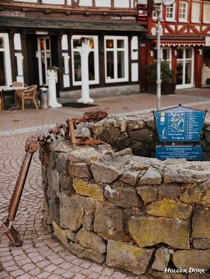 Ein historischer Ziehbrunnen aus groben Natursteinen auf dem Marktplatz in Grünberg. Über den Rand des Brunnens lehnt eine rostige Metallskulptur, der „Brunnengucker“, die einen menschenähnlichen Oberkörper mit Rippen, Armen und einem Kopf darstellt. Die Figur scheint in den Brunnen zu schauen. Neben dem Brunnen befindet sich eine Infotafel mit einer Karte des historischen Rundgangs von Grünberg. Im Hintergrund sind Fachwerkhäuser mit weißen Fenstern und gemütlicher Außengastronomie zu sehen.