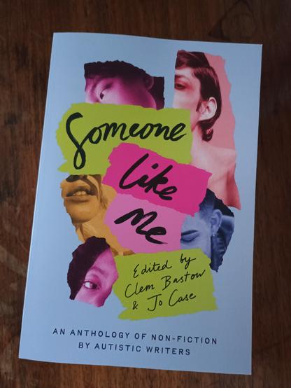 The front cover of the anthology Some Like Me. Its background colour is pale purple and the title is printed over slices of yellow and pink on top of images showing people ripped into thin slithers collage style. The book is sitting on a wooden surface.