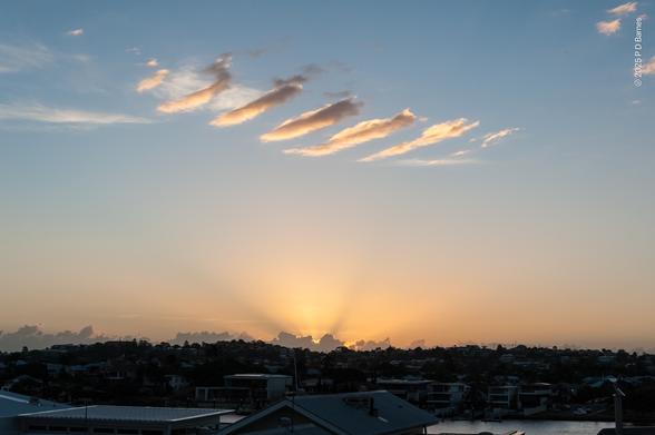 A sunrise over a suburban ridgeline, with a series of small, linear, parallel clouds high in the sky, looking like a barcode.