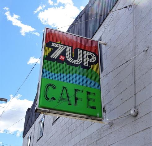Vintage plastic 7-Up sign attached to a cinder block building. The top portion says 7-UP in letter made of white dots with a rainbow background also made of dots. The bottom portion says Cafe in faded black block letters with a bright green background.