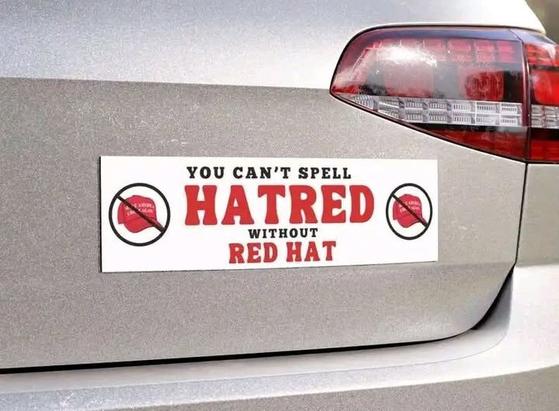 Bumper sticker on the back of a car. The sticker reads: "YOU CAN'T SPELL HATRED WITHOUT RED HAT." The words "HATRED" and "RED HAT" are in bold red letters. There are two symbols on either side of the text, each showing a red hat with a diagonal line through it, indicating a prohibition.