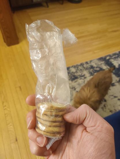 Three peanut butter sandwich cookies left in a plastic sleeve. A wood floor and a small brown dog are in the background.
