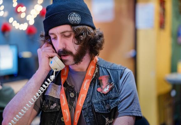 A man with a mutton-chop beard and a septum ornament, sporting a wool cap and a denim vest with flair, appears to be speaking on a classic wired telephone receiver.