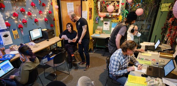 We see, largely from above, the WFMU Fundraising Marathon phone room, with station manager Ken Freedman at the emcee microphone and volunteers arrayed at tables around the room staffing telephones and computers.