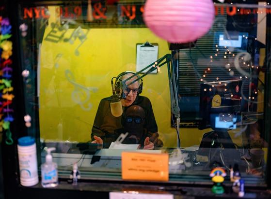 Looking through a glass window framed with brightly-colored decorations,  we see a man (DJ Dave Mandl) speaking into a radio microphone on an articulating microphone stand arm.