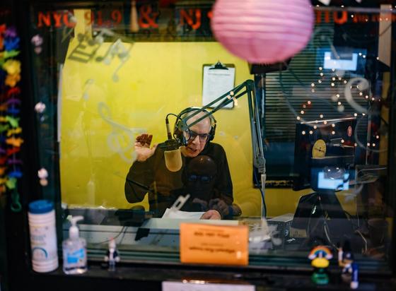 Looking through a glass window framed with brightly-colored decorations,  we see a man (DJ Dave Mandl) speaking into a radio microphone on an articulating microphone stand arm.  Mr. Mandl has his right hand raised to gesticulate.