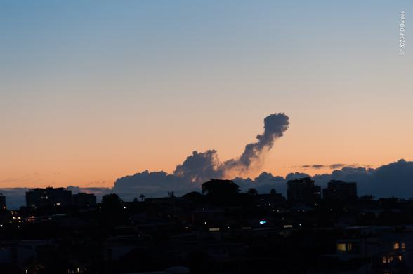 Dark suburban horizon at dawn, with clouds immediately behind. One cloud has formed into the rough outline of an elephant with an upraised trunk, possibly blowing smoke. The sky behind is peach, grading to blue at the top.