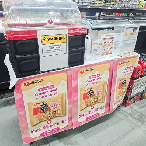 Photo of a free sample booth at an Asian-American supermarket. The booth is set up to prepare and give out little cups of Spicy & Creamy Toomba Stir Fry Style Noodles with Sauce Mix from Shin Ramyun.