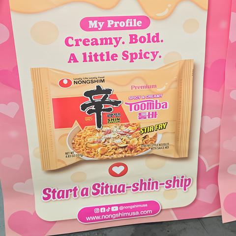 Photo of a promo poster for Spicy & Creamy Toomba Stir Fry Style Noodles with Sauce Mix from Shin Ramyun taped to the front of a free sample booth at an Asian-American supermarket. The poster predominantly features the text "Start a Situa-shin-ship".

A basic transcript of the poster follows.

My Profile

Creamy. Bold. A little Spicy.

Start a Situa-shin-ship.