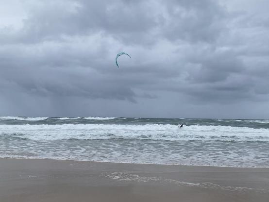 Kite surfer riding the white waves under grey cloudy skies, light blue kite sail full of air. Taken from the sandy beach looking out toward the ocean horizon.