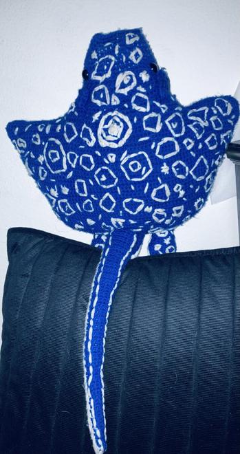 A top shot of the plush showing the full length of the tail which is also embroidered to mimic a spotted eagle ray. You can also see fin like appendages attached to the ray, which are also embroidered to match a spotted eagle ray. The plush is propped on a black pillow.